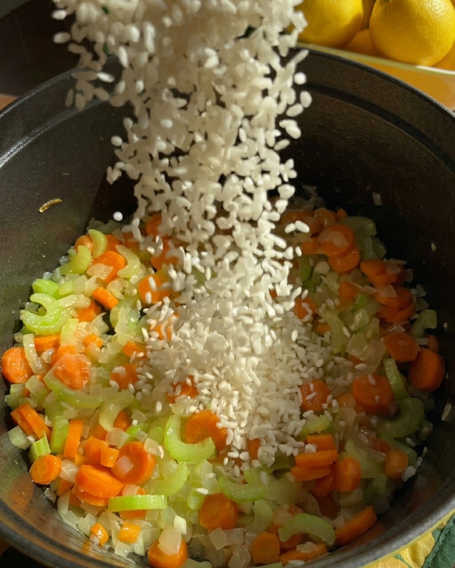 Un chorro de arroz jazmín blanco se vierte en una olla oscura llena de zanahorias, apio y cebollas picadas y sofritas.
