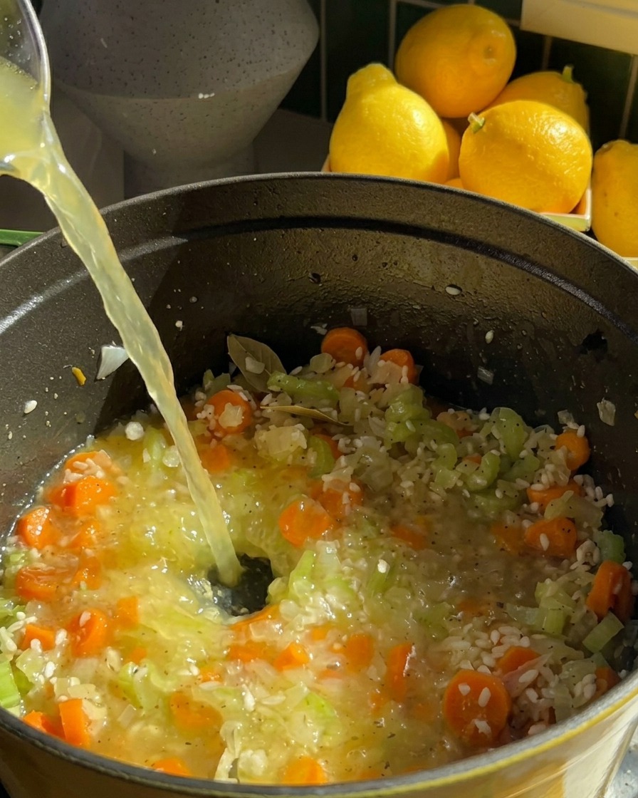 Un chorro de caldo de pollo transparente se vierte en una olla que contiene arroz, zanahorias y apio.