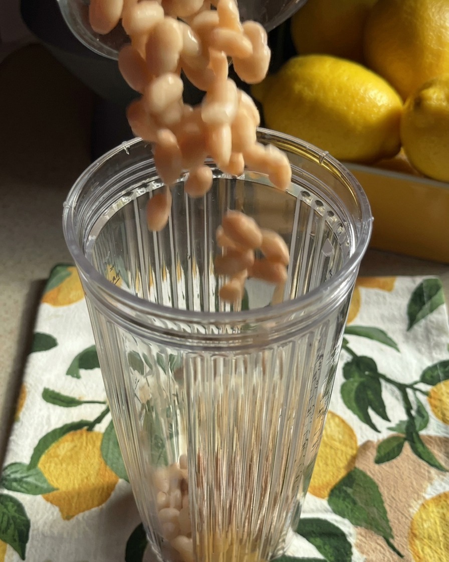 Alubias blancas cocidas caen en el vaso transparente de una batidora situado sobre un paño de cocina con estampado de limones.