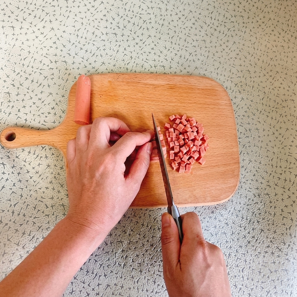 Unas manos usando un cuchillo de chef para cortar una salchicha de jamón rosada en cubos pequeños y uniformes sobre una tabla de madera clara.