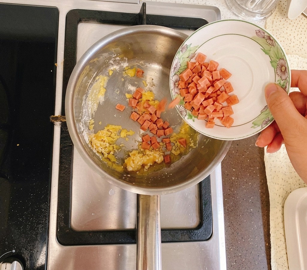 Jamón en cubos siendo vertido desde un pequeño plato blanco en una olla que contiene una mezcla dorada de mantequilla y harina.