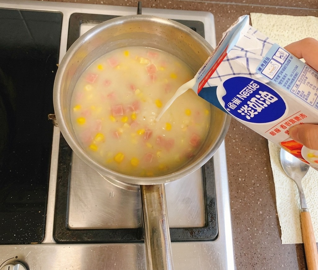 Crema ligera siendo vertida desde un cartón azul y blanco en una olla de sopa de maíz cremosa que hierve a fuego lento en la estufa.