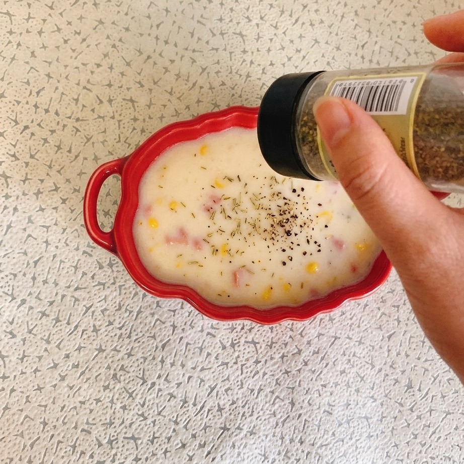 Una mano sosteniendo un frasco de especias y espolvoreando hierbas secas sobre un tazón rojo de sopa cremosa de maíz con jamón.