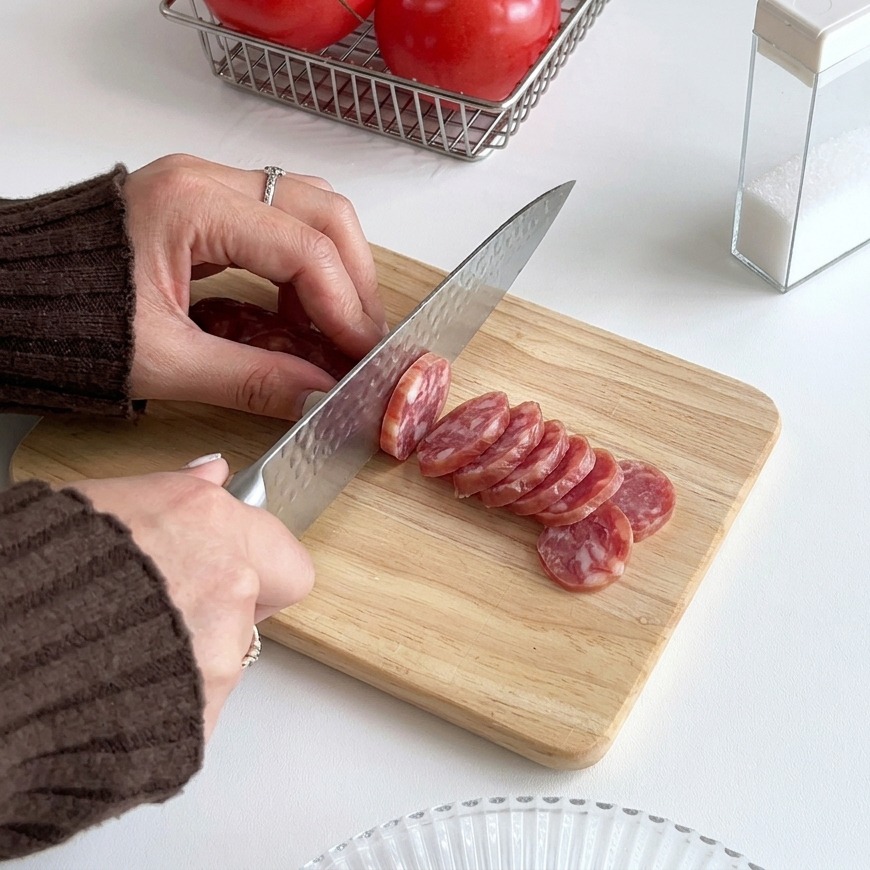 Manos usando un cuchillo de cocina plateado para cortar una salchicha roja en rodajas finas sobre una tabla de cortar de madera clara.