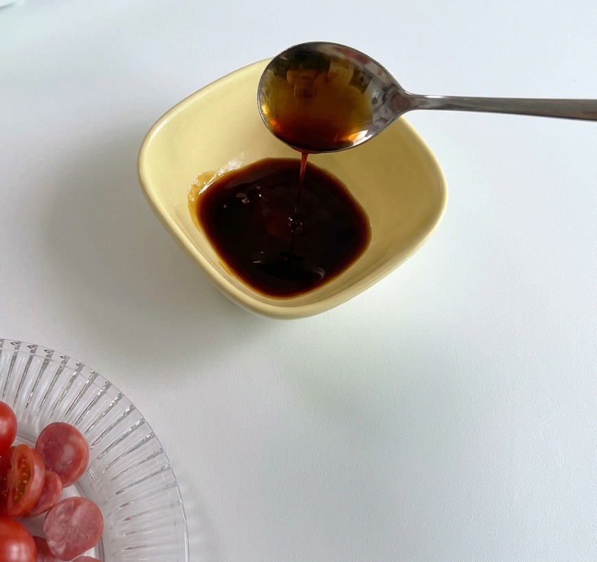 Una cuchara de metal vertiendo salsa de soja oscura en un tazón amarillo de preparación que ya contiene una porción de salsa de tomate roja.