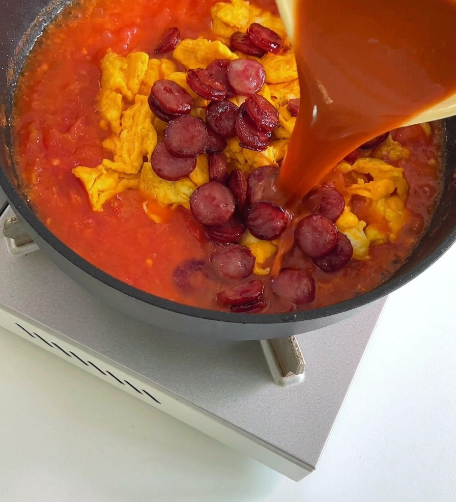 Una salsa rojiza vertiéndose desde un tazón amarillo en una sartén que contiene huevos revueltos y salchichas cortadas en una mezcla a base de tomate.