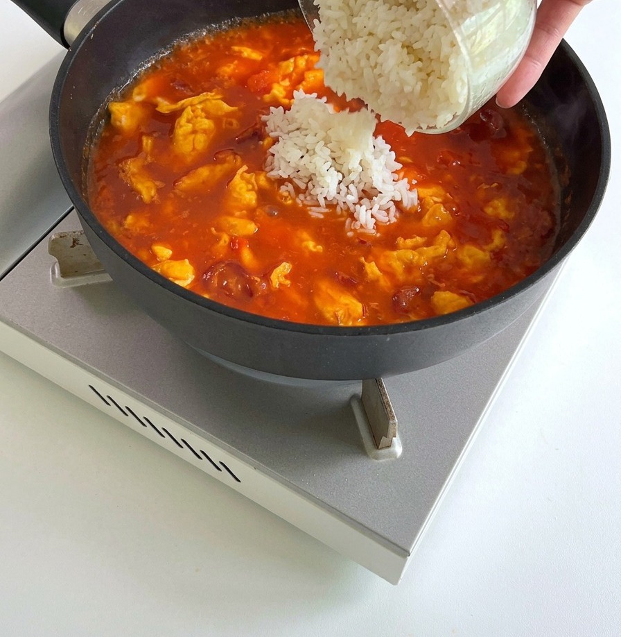 Arroz blanco cocido volcándose desde un tazón de vidrio en una sartén con salsa de tomate burbujeante, huevos y salchichas.