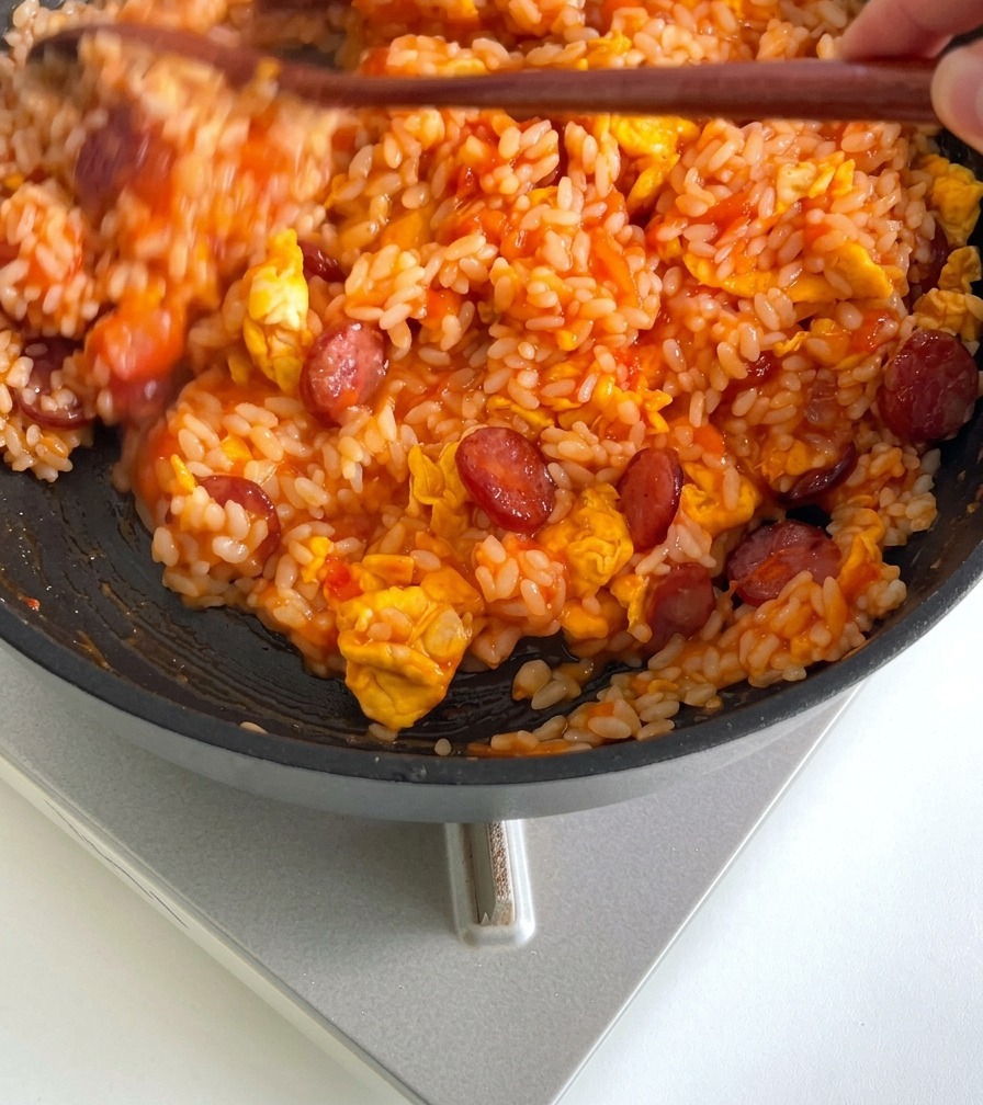 Arroz mezclándose con un utensilio de cocina de madera en una sartén con salsa de tomate, salchichas y huevos hasta integrarse por completo.