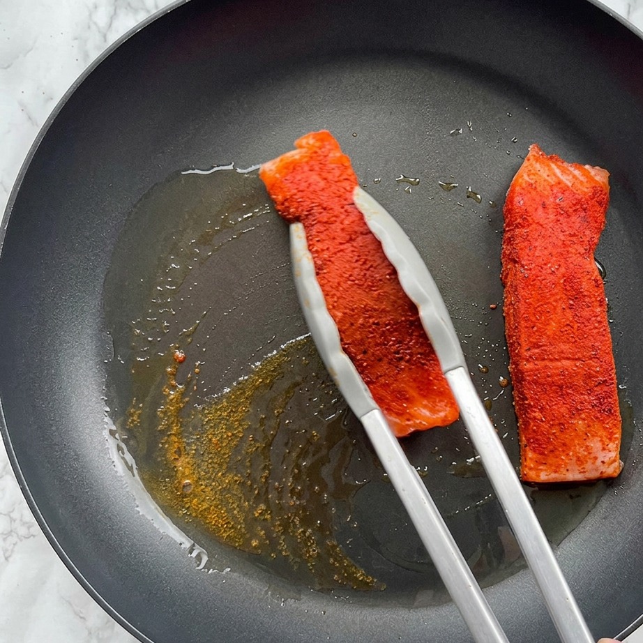 Un filete de salmón sazonado dorándose en una sartén caliente con aceite de oliva burbujeante, sostenido por unas pinzas de metal.