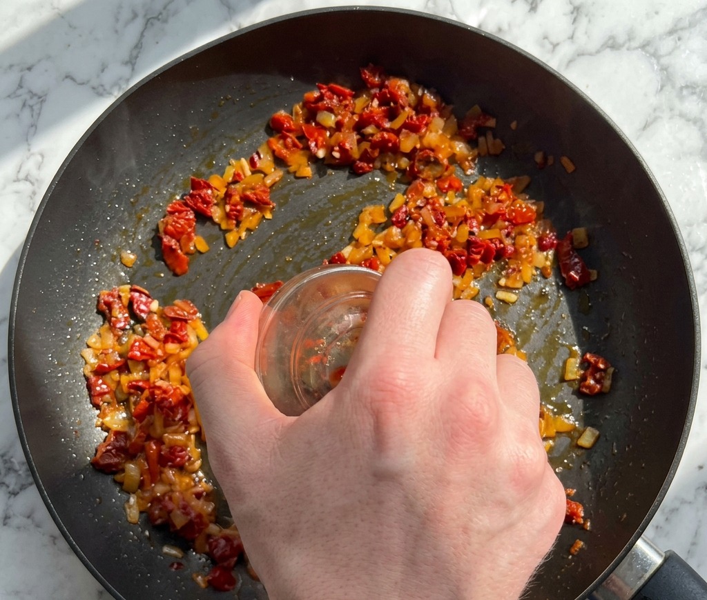 Una mano vertiendo tomates secos picados y hojuelas de pimiento rojo desde un bol de vidrio a la sartén con el sofrito.