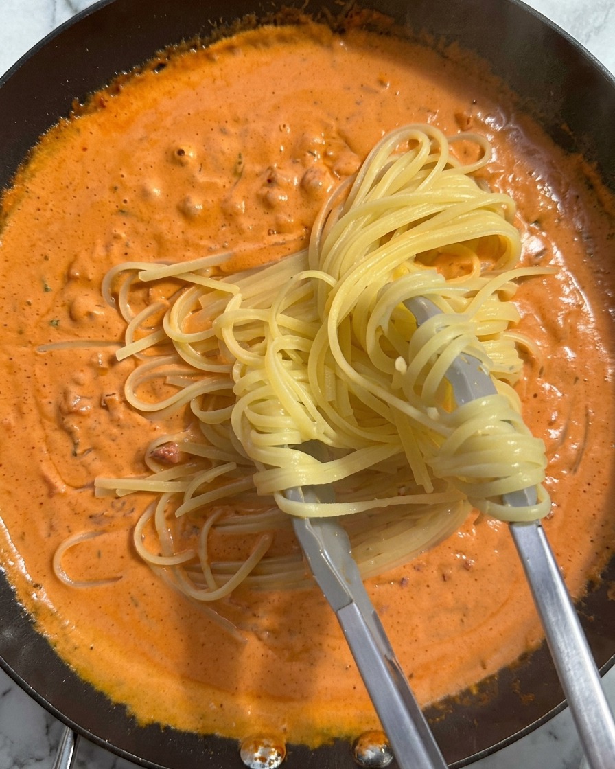 Una porción de pasta cocida siendo levantada con pinzas e introducida en una sartén con una rica salsa naranja.
