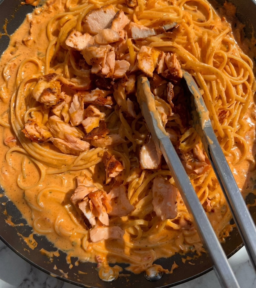 Trozos de salmón rosado cocido siendo mezclados en una sartén de pasta cremosa usando pinzas de metal.
