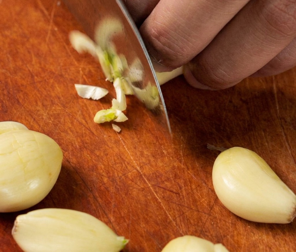 Primer plano de un cuchillo de chef picando finamente varios dientes de ajo fresco sobre una tabla de cortar de madera.