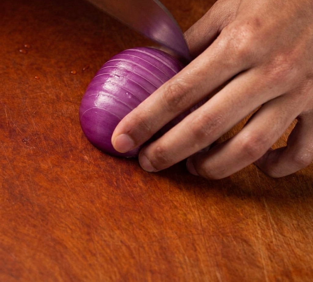 Un cuchillo afilado cortando una cebolla morada en cubos finos y uniformes sobre una tabla de madera.