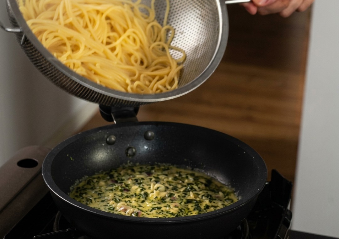 Espaguetis cocidos vertiéndose desde un colador de acero inoxidable en una sartén con salsa cremosa salpicada de albahaca verde.