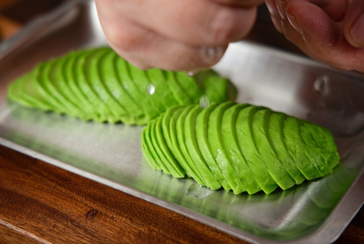 Zumo de limón fresco siendo exprimido a mano sobre una bandeja de láminas de aguacate perfectamente dispuestas.