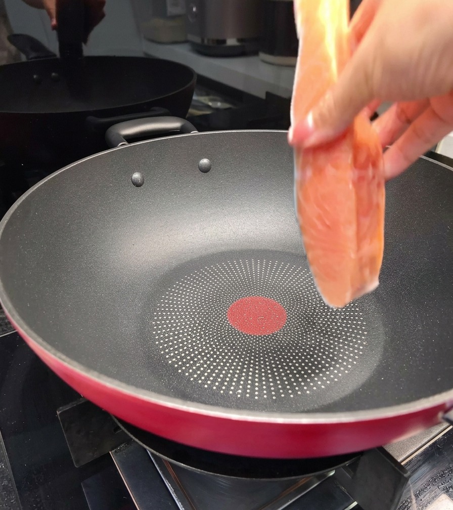 Una mano colocando un filete de salmón fresco en una sartén antiadherente negra grande sobre la estufa.