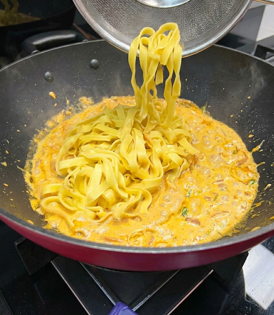 Fideos anchos de fettuccine siendo levantados con un colador de malla e incorporados a una sartén con salsa cremosa naranja que hierve a fuego lento.