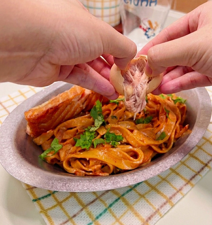 Manos pelando y colocando con cuidado un huevo tibio sobre un plato servido con pasta cremosa picante y salmón dorado.