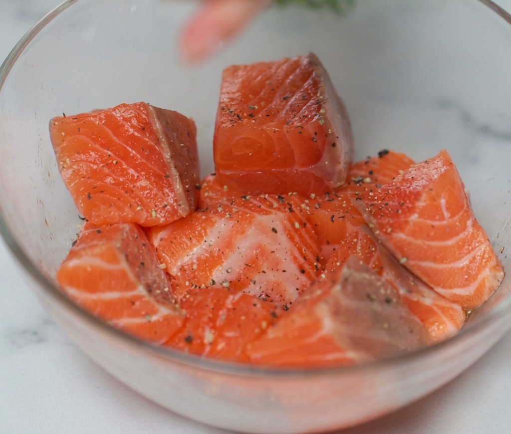 Dados de salmón crudo en un tazón de vidrio siendo espolvoreados con sal y pimienta negra de un molinillo.