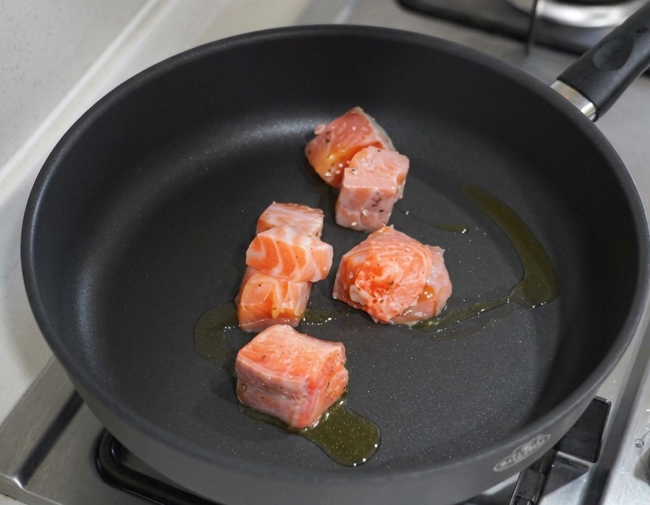 Trozos de salmón en dados dorándose en una sartén negra antiadherente con aceite de oliva sobre una estufa de gas.