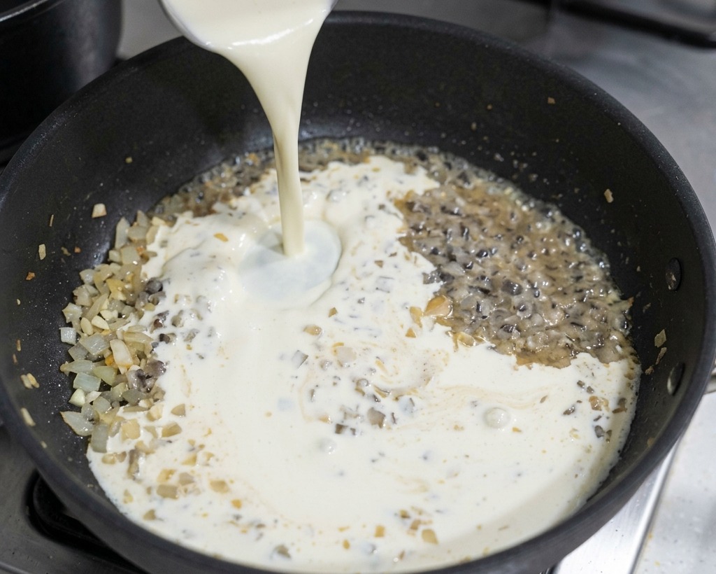 Crema de leche siendo vertida en una sartén negra que contiene cebollas sofritas y vino para crear una salsa blanca.