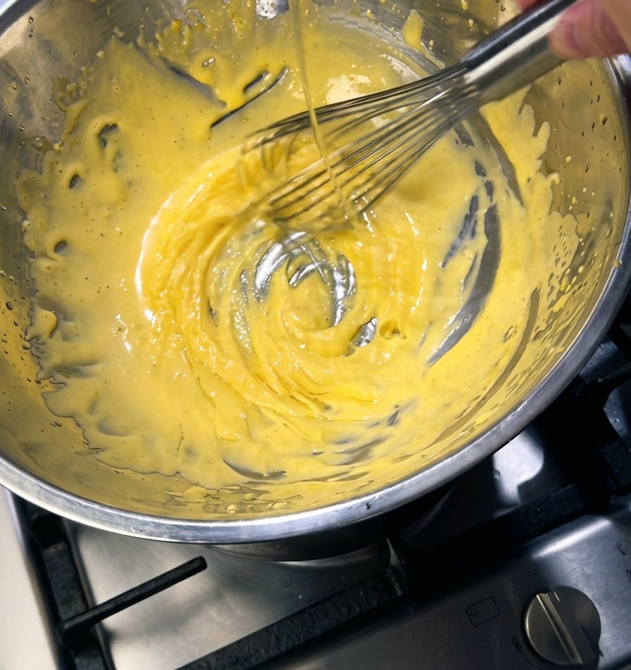 Un batidor de metal removiendo una mezcla espesa y de color amarillo pálido en un bol de acero inoxidable mientras se vierte un fino hilo de mantequilla dorada.
