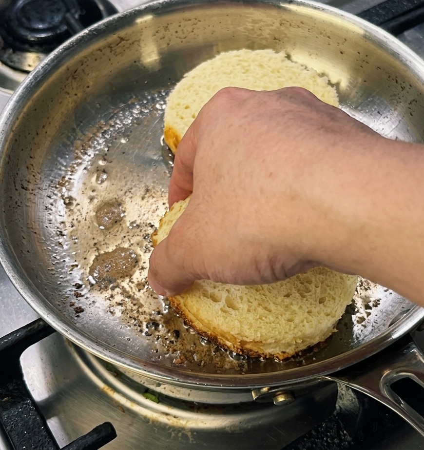 Una mano presionando dos rebanadas gruesas y redondas de pan brioche en una sartén para lograr una corteza dorada.