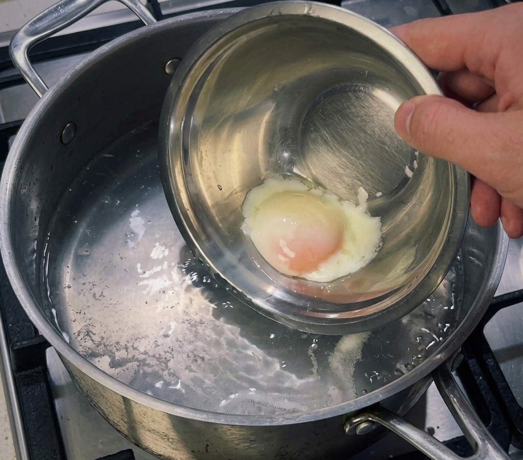 Un pequeño cuenco de metal que contiene un huevo cocinado a baja temperatura sumergiéndose en un cazo de agua caliente para reafirmar la clara.