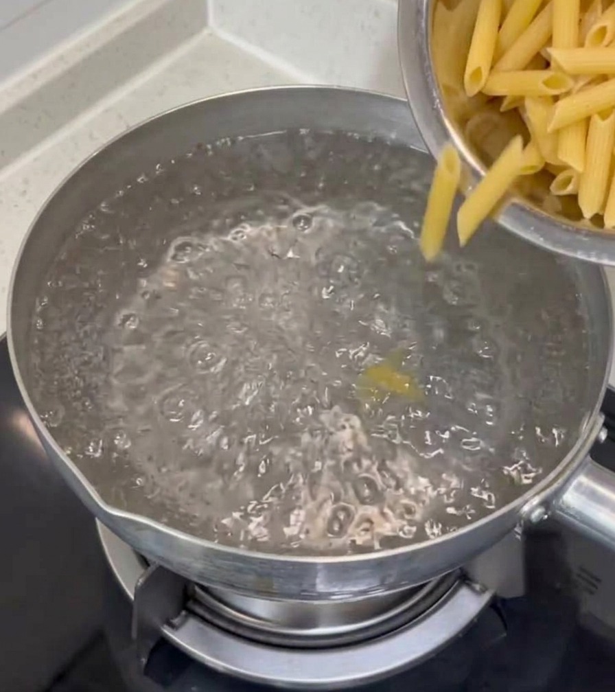 Pasta penne seca vertiéndose desde un tazón de acero inoxidable a una olla con agua hirviendo sobre la estufa.