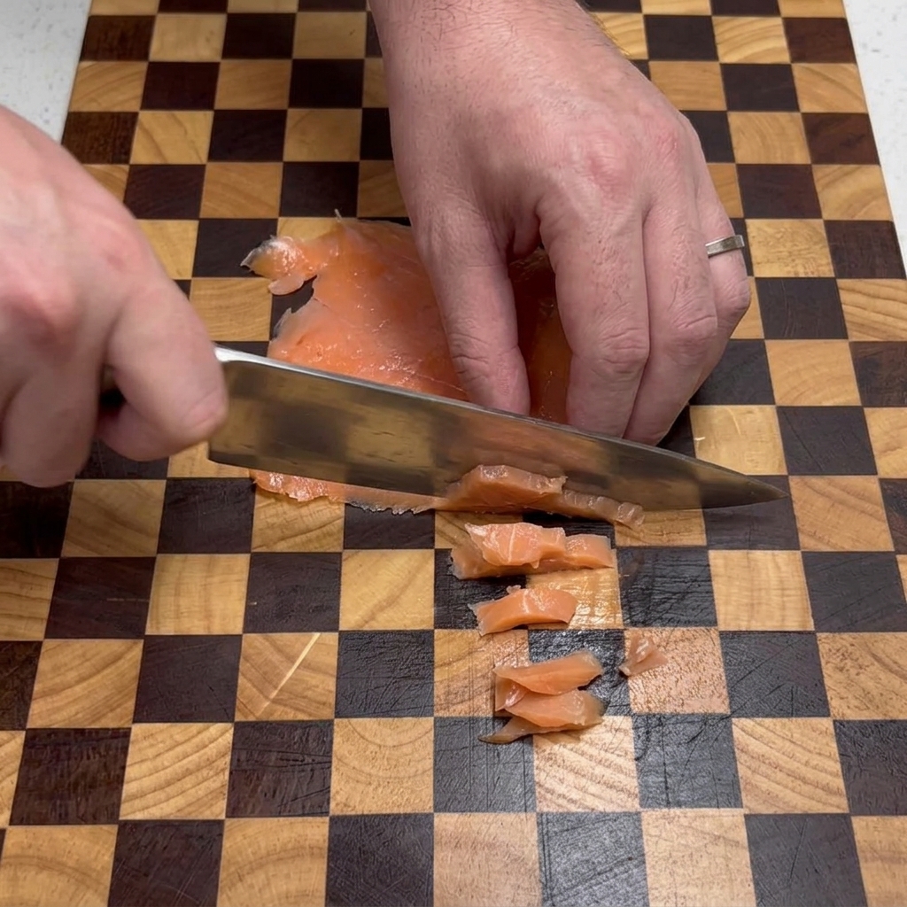 Manos de chef usando un cuchillo plateado para cortar trozos planos de salmón ahumado en pequeños cuadrados sobre una tabla de cortar.