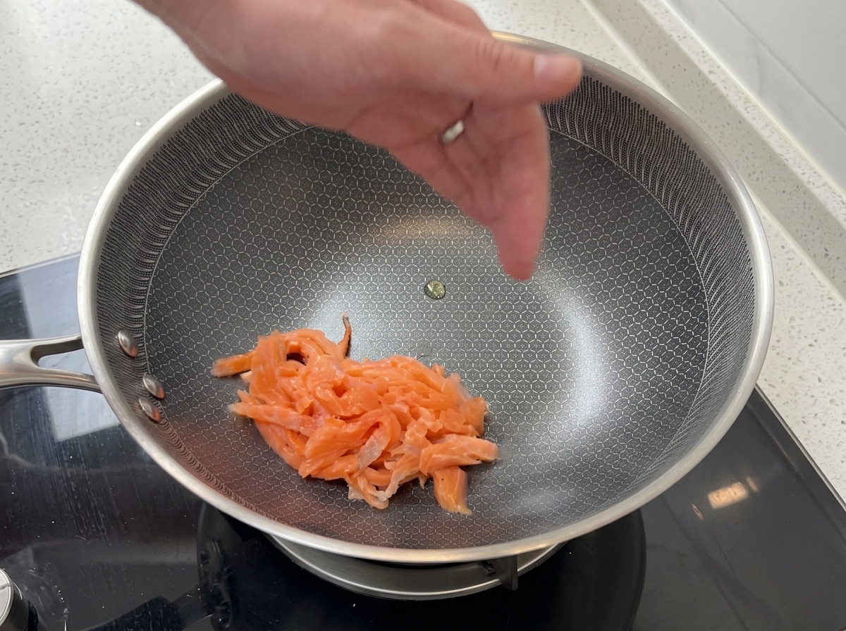 Rebanadas de salmón ahumado rosado salteándose en una sartén de acero inoxidable con un patrón hexagonal texturizado.