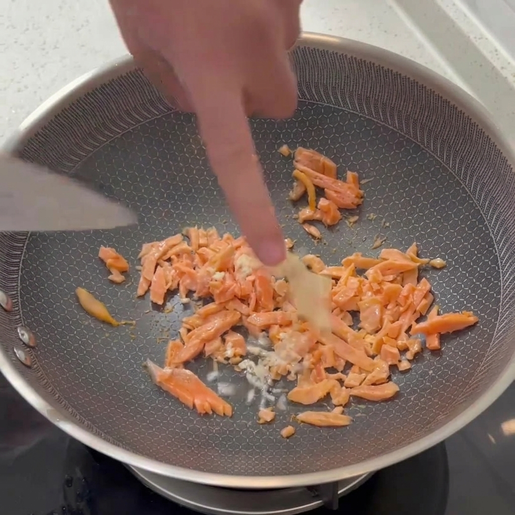 Ajo blanco picado mezclándose con una espátula en una sartén con trozos de salmón ahumado parcialmente cocidos.