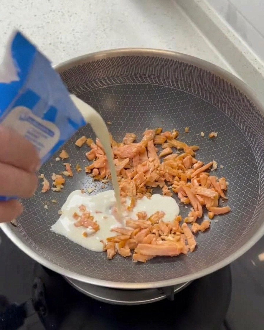 Una mano vertiendo crema de leche blanca desde un cartón azul y blanco en una sartén que contiene trozos de salmón cocido.