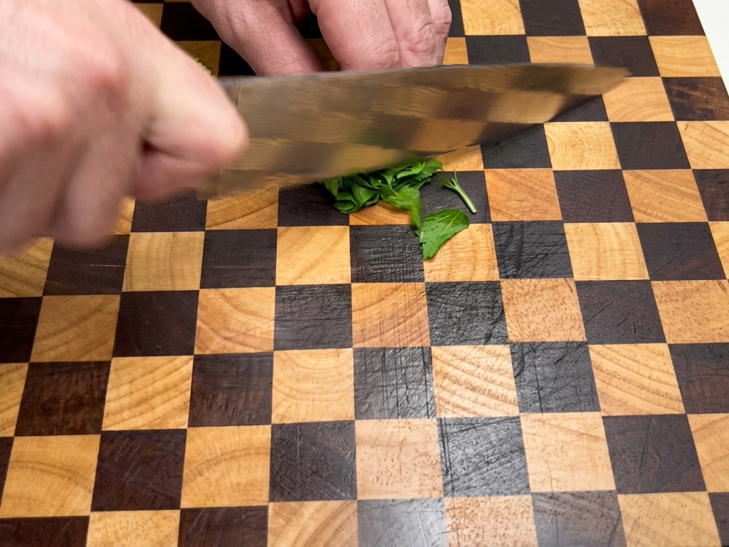 Primer plano de manos usando un cuchillo de chef para picar finamente hojas de perejil verde fresco sobre una tabla de cortar de madera.