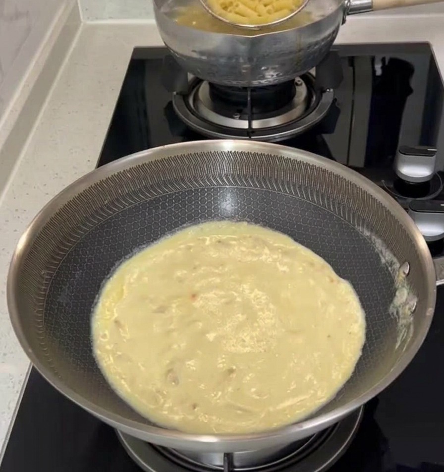 Un colador de araña de metal levantando pasta penne cocida de una olla hirviendo y pasándola a una sartén llena de salsa cremosa blanca en la estufa.