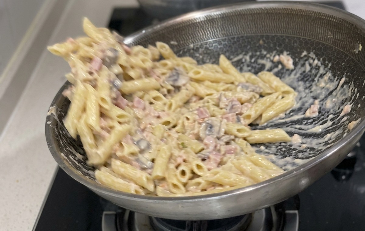 Pasta penne cocida mezclándose con una salsa blanca cremosa y trozos de salmón en una sartén grande de acero inoxidable.