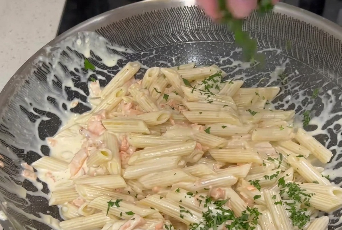 Una mano espolvoreando perejil verde finamente picado sobre una sartén grande de pasta penne mezclada con salsa blanca cremosa y trozos de salmón ahumado rosado.
