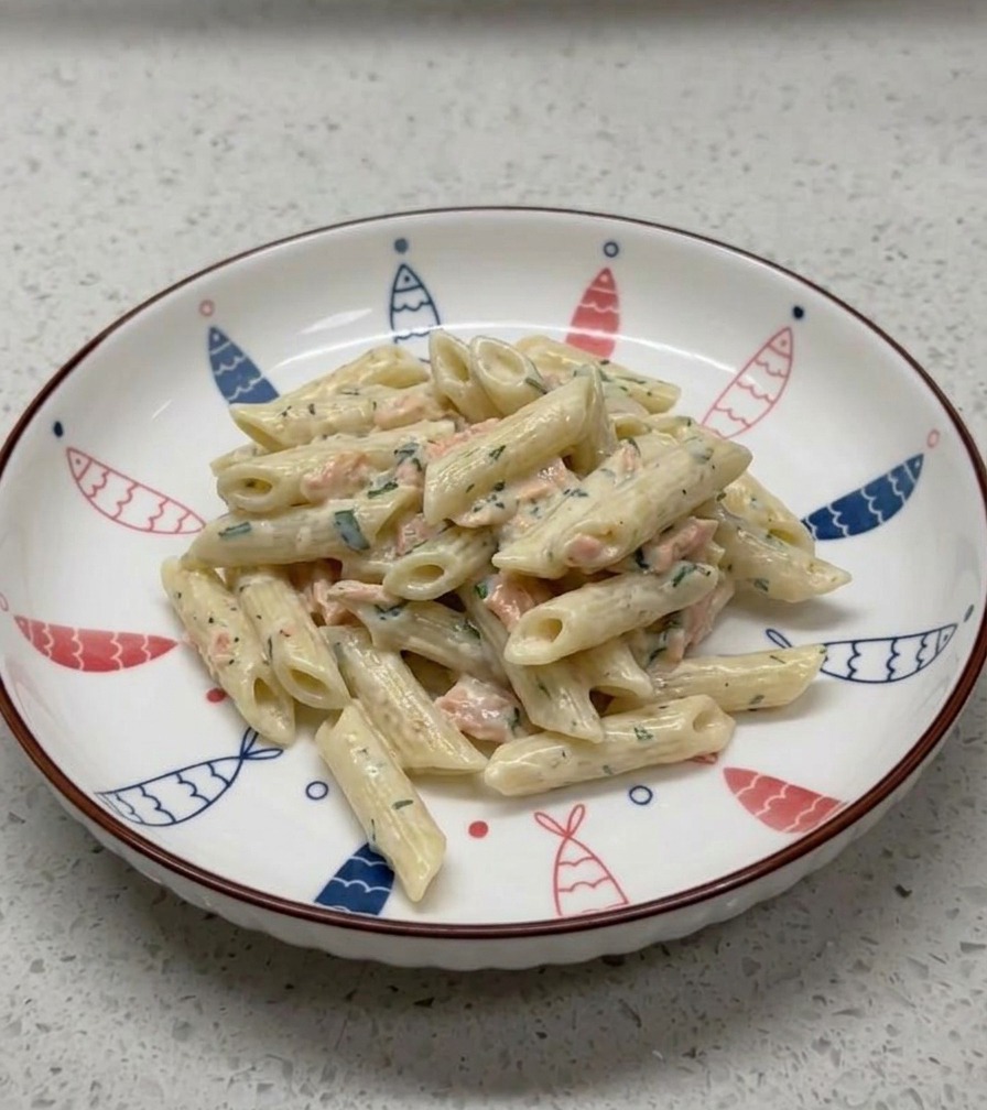 Una porción de pasta penne cremosa con salmón servida cuidadosamente en un tazón de cerámica blanca decorado con un patrón de peces azules y rojos alrededor del borde.