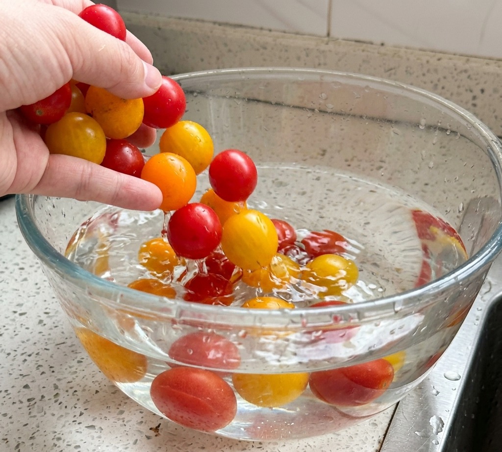Una mano lavando tomates cherry rojos y amarillos en un tazón grande de vidrio lleno de agua.