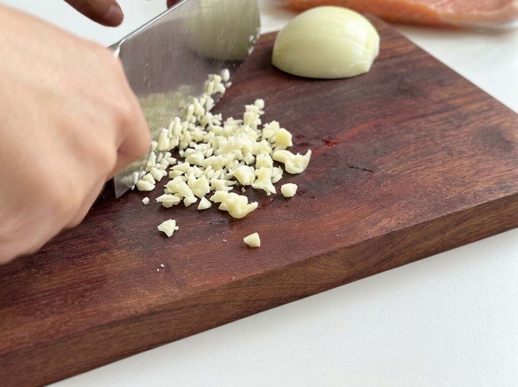 Primer plano de una persona usando un cuchillo de chef para picar finamente cebolla blanca y ajo sobre una tabla de madera.