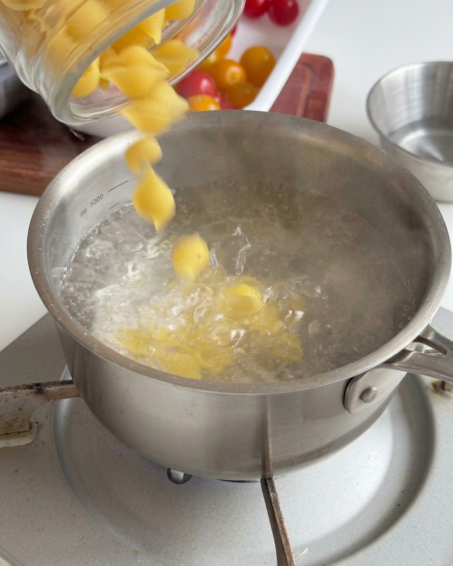 Pasta amarilla con forma de caracola vertiéndose desde un frasco de vidrio en una olla de acero inoxidable con agua hirviendo.