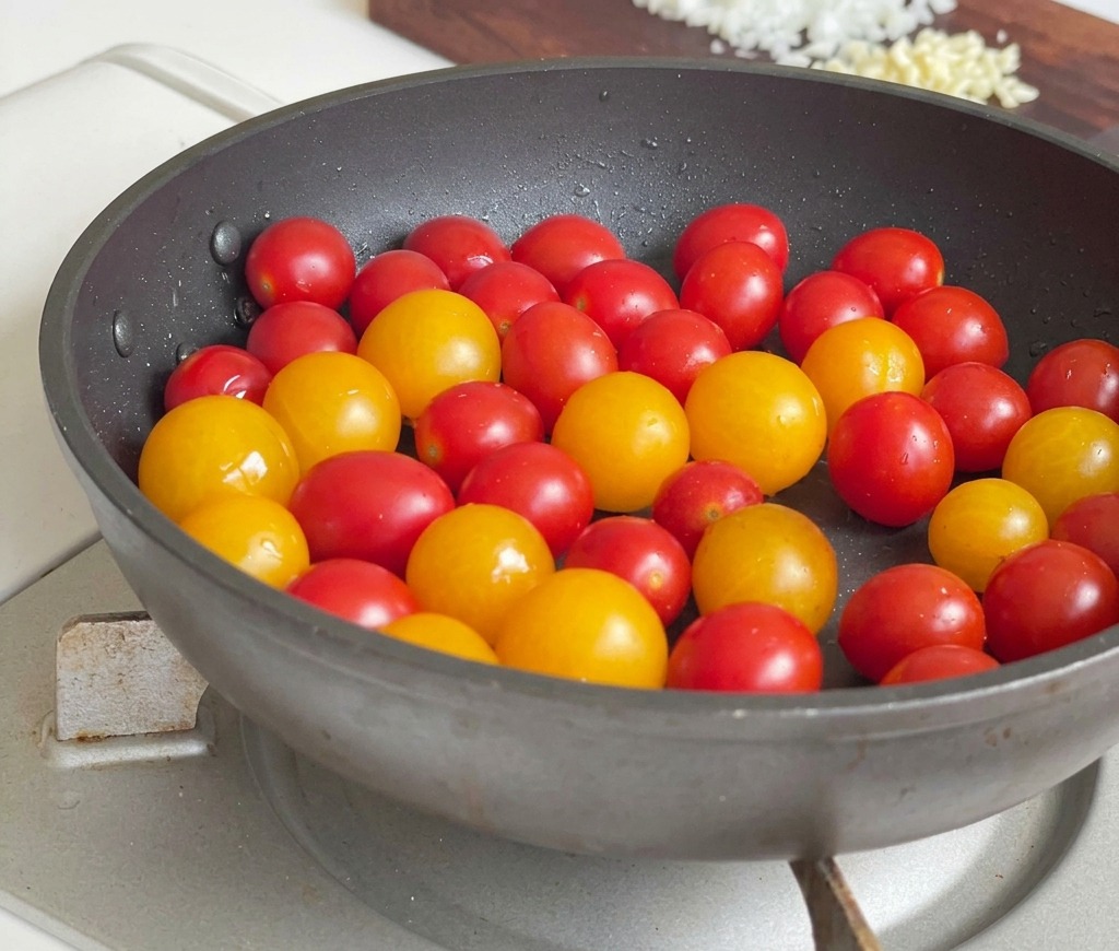 Una mezcla de tomates cherry rojos y amarillos salteándose en una sartén oscura sobre una estufa de gas.