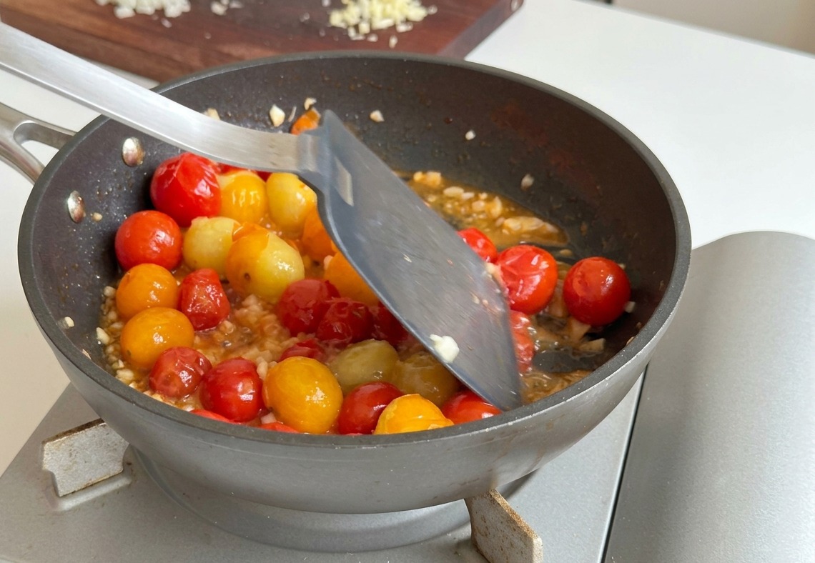 Ajo y cebolla picados mezclándose con una espátula metálica en una sartén con tomates cherry ya blandos.