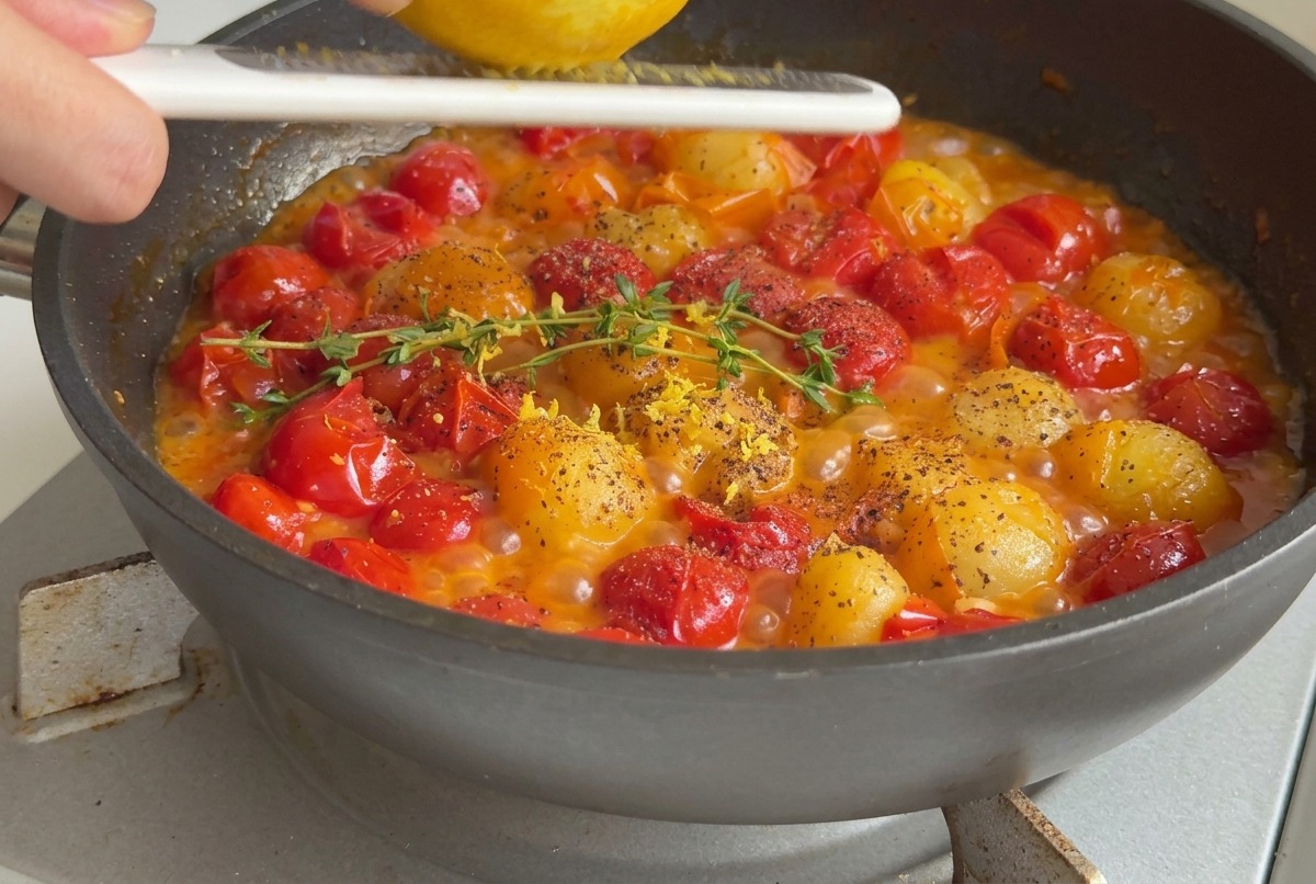 Una mano rallando cáscara de limón sobre una sartén con tomates cherry hirviendo sazonados con tomillo fresco y pimienta negra.