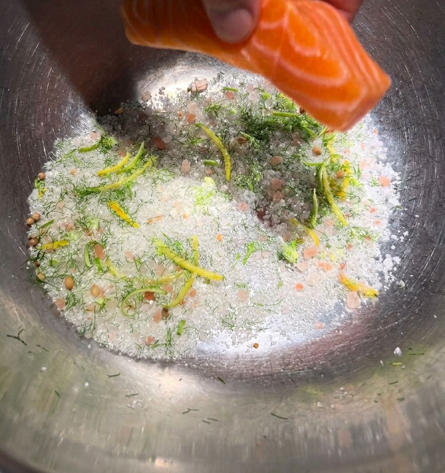 Una mano colocando un filete de salmón de color naranja vibrante sobre una cama de sal de curado y hierbas verdes en un bol de metal.
