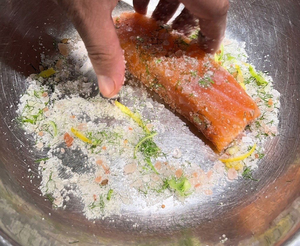 Manos frotando una mezcla gruesa de sal, azúcar y hierbas sobre un trozo grueso de salmón crudo en un bol de metal.