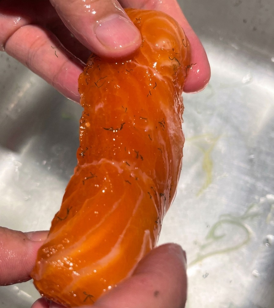 Una mano sosteniendo un filete de salmón curado bajo un chorro de agua corriente para quitar la mezcla de sal y especias.