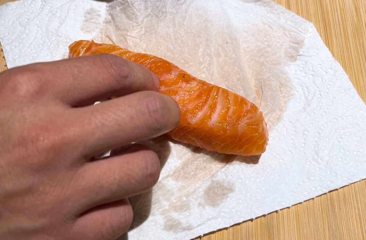Una mano usando una toalla de papel blanca para secar a toques un filete de salmón curado de color naranja brillante sobre una tabla de madera.