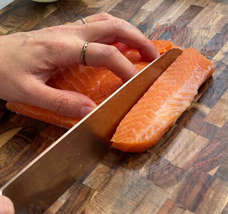 Primer plano de unas manos usando un cuchillo para cortar un filete grande de salmón crudo en dados pequeños sobre una tabla de madera.